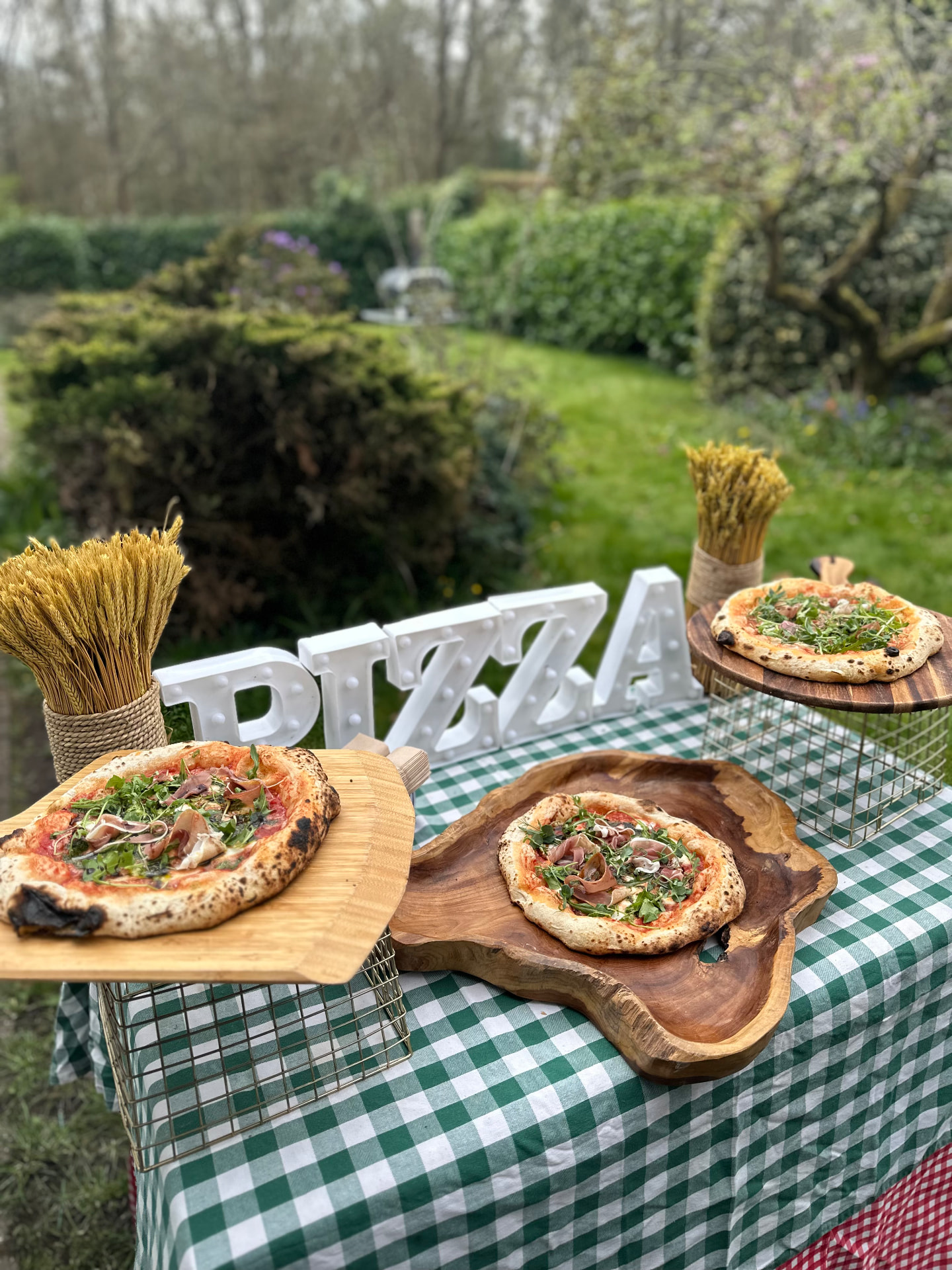 Sourdough pizzas on boards at a garden pizza table with a lit PIZZA sign.