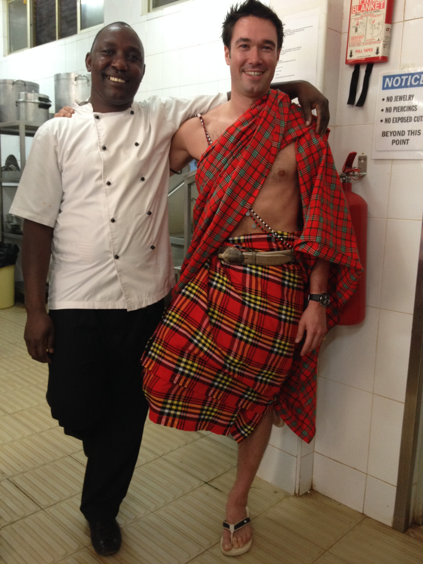 Gregory in a Maasai shuka, arm in arm with a chef in a Kenyan kitchen.