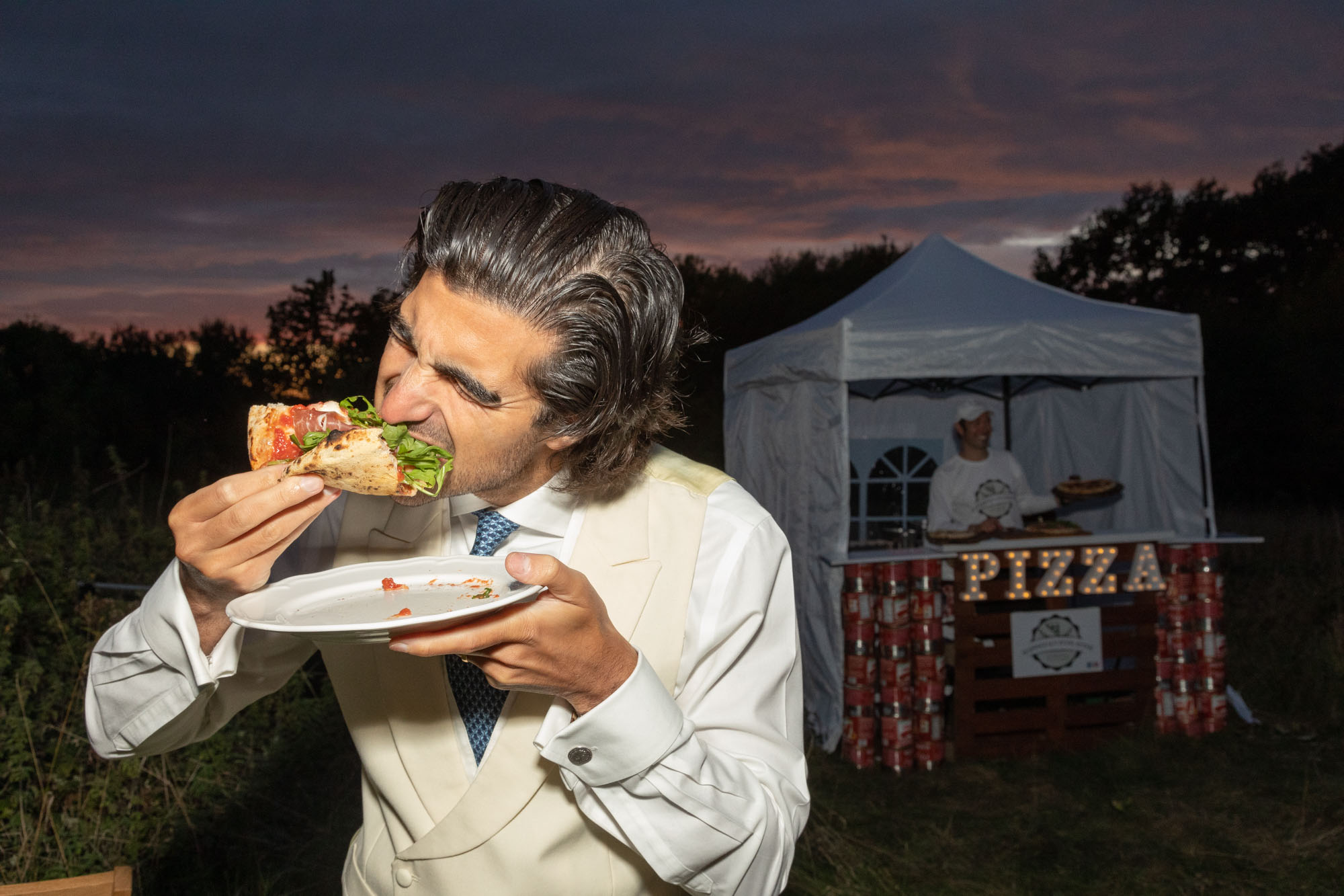 A guest in a cream suit biting into a slice in front of the pizza stand.