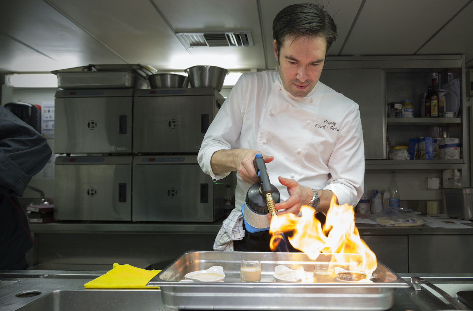 Gregory using a blowtorch over a tray of desserts in a professional kitchen.