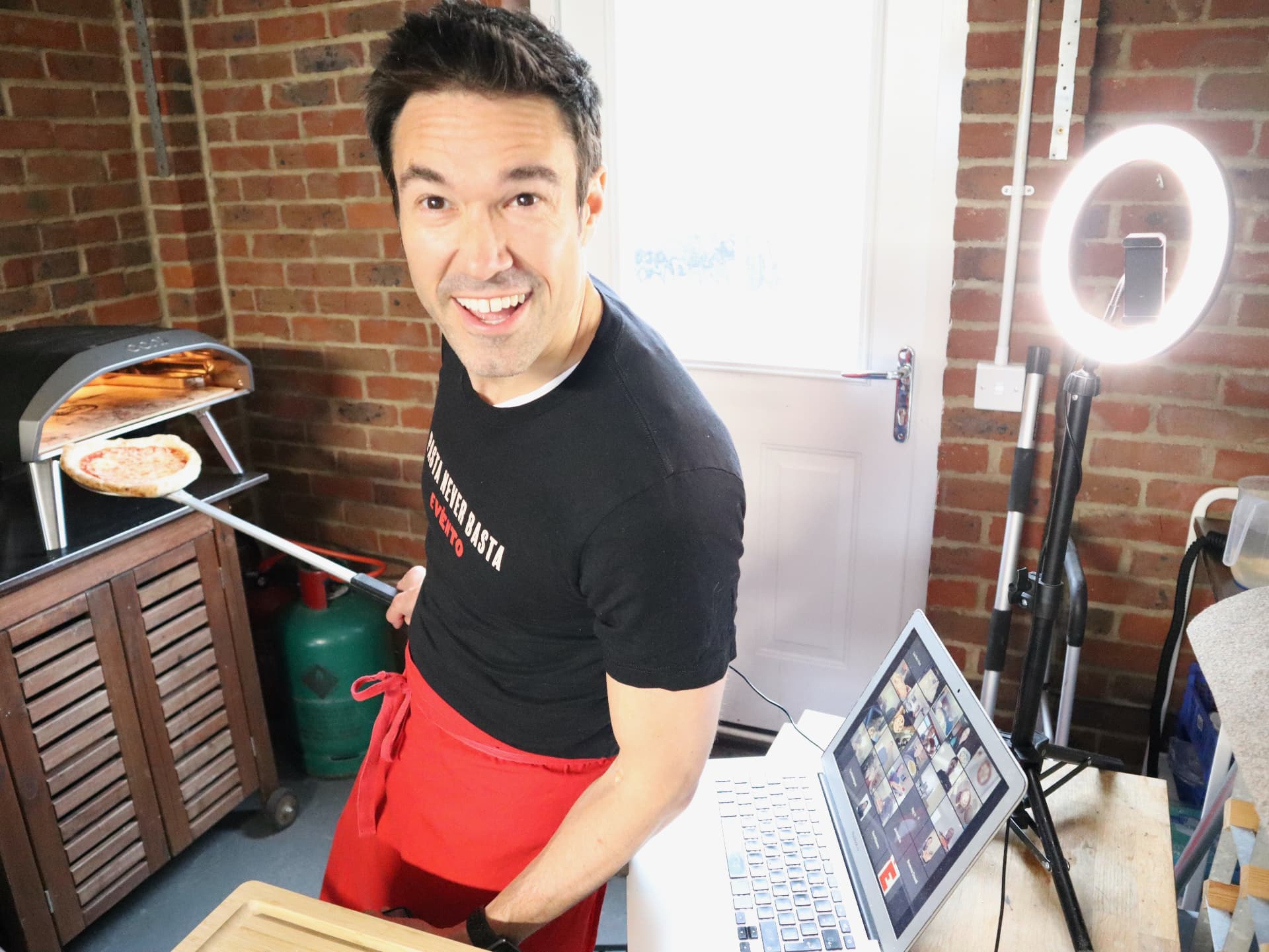 Gregory laughing beside a pizza oven while hosting a live online class.