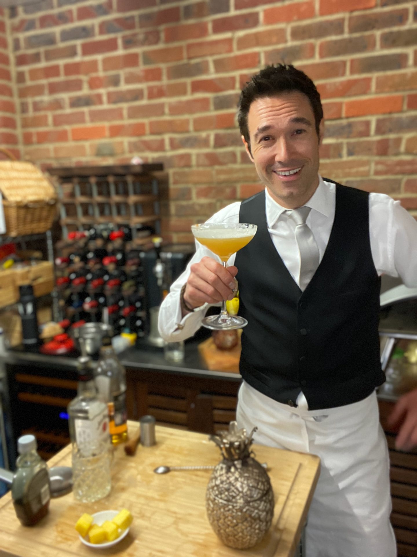 Gregory in a waistcoat and tie with a cocktail in a brick-walled bar.