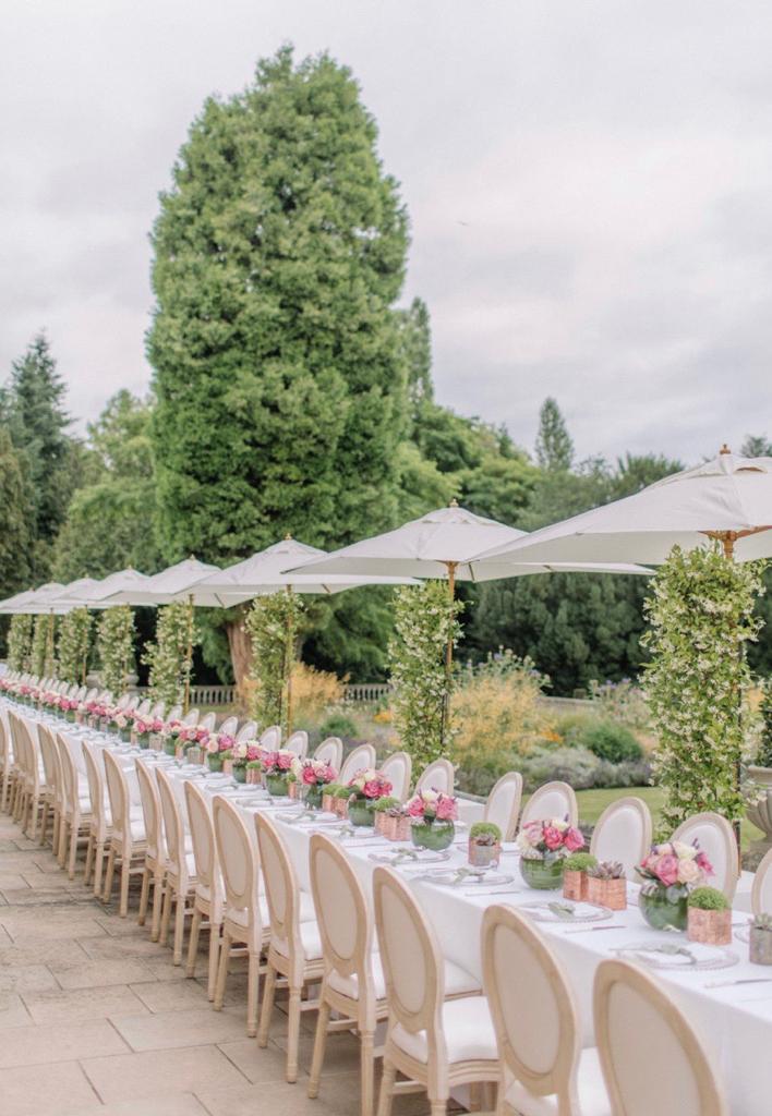 A long garden dining table with pink flowers and white parasols.