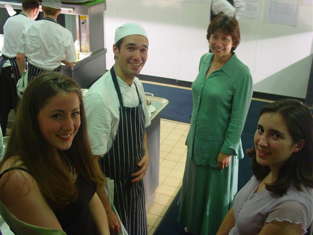 Gregory as a young commis chef in a striped apron with three guests in a kitchen.